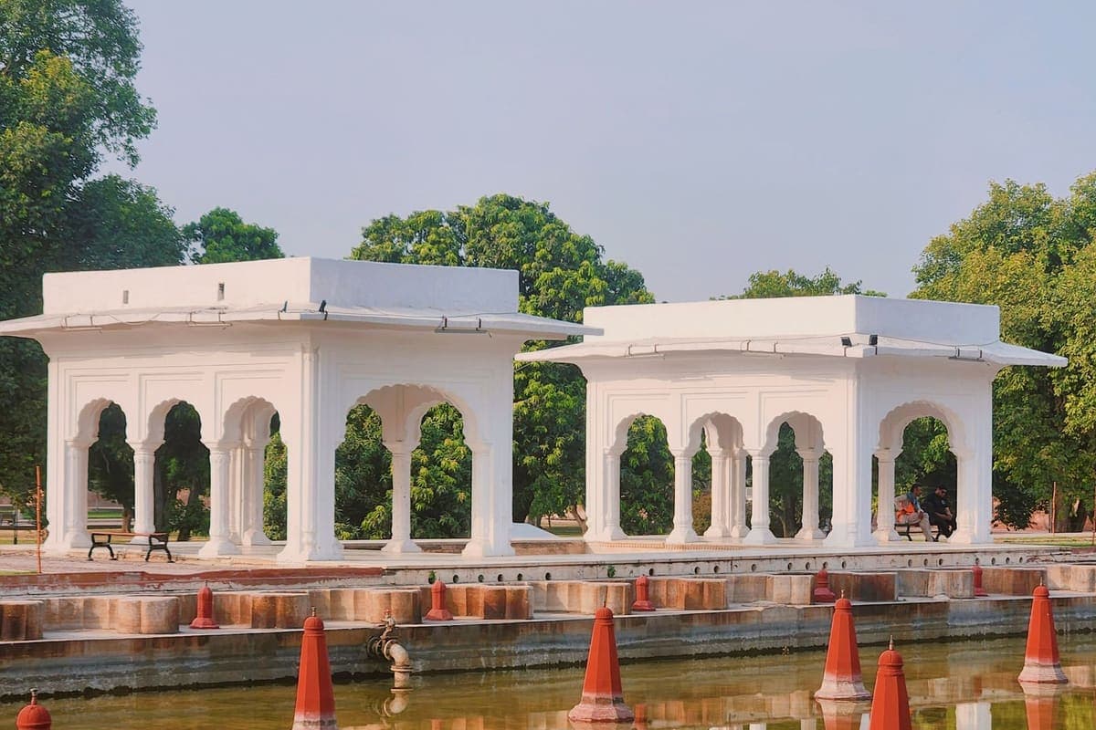 A white Mughal-style garden pavilion with red pillars, mirrored in a still rectangular water channel